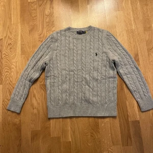 Grå stickad tröja från Polo Ralph Lauren - Säljer en snygg grå stickad tröja från Polo Ralph Lauren med klassiskt kabelstickat mönster och rund hals. Tröjan har långärmade ribbade muddar och den ikoniska Polo-loggan broderad på bröstet. Perfekt för dig som gillar stilren och tidlös design.