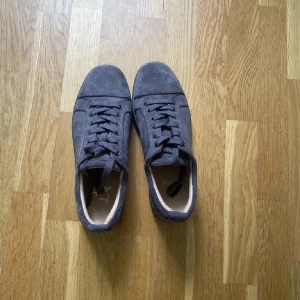 Helt nya oanvändas Grå mockasneakers från Christian Louboutin - Snygga sneakers i grå mocka från Christian Louboutin med klassisk röd sula. Skorna har rund tå, platt sula och snörning. Perfekta för dig som vill ha en lyxig och stilren look med en ikonisk detalj under foten.