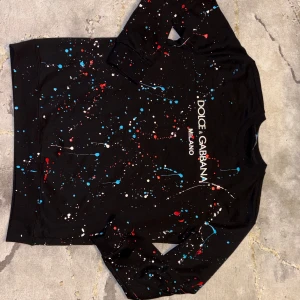 Dolce & Gabbana Svart Tröja - Svart sweatshirt från Dolce & Gabbana med coolt färgstänksmönster i rött, blått och vitt. Tröjan har rund hals, långa ärmar och tryckt logga framtill. Perfekt för dig som vill sticka ut med en unik och trendig look