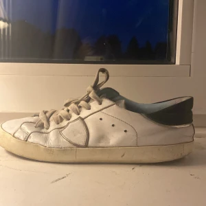 Vita Philippe Model sneakers  - Snygga vita sneakers från Philippe Model Paris med svarta detaljer på hälen och beige snörning. Skorna är i skinn och har en klassisk låg profil med rund tå och platt sula. Perfekta för dig som gillar stilrena och trendiga sneakers.