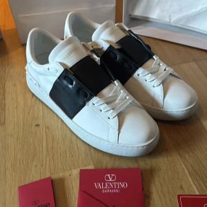 Valentino Garavani Open sneakers vit/svart - Säljer ett par Valentino Garavani Open sneakers i vit skinn med bred svart läderdetalj över mitten. Klassisk rund tå, platt sula och vita snören. Märkeslogga på plösen och insidan. Kommer med originalkartong och dustbag.
