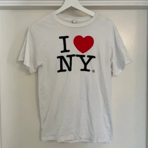 Vit I ❤️ NY t-shirt - Vit klassisk t-shirt med det ikoniska I ❤️ NY-trycket på bröstet. T-shirten har rund halsringning och korta ärmar. Perfekt för dig som vill visa lite kärlek till New York och gillar en enkel, clean stil.