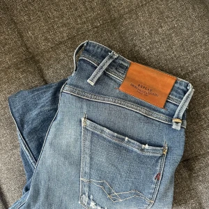 Replay anbass blå slim jeans med slitningar - Säljer ett par blå slim jeans från Replay med snygga slitningar och ljusare tvätt på låren. Klassisk femficksmodell med läderpatch bak i midjan och detaljerade bakfickor. Jeansen är i stretchigt denim och har en modern, slim passform. De är i bra skick.