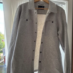 Grå overshirt från Pelle P - Snygg grå overshirt från Pelle P med klassisk krage, tryckknappar och två bröstfickor. Mjuk och skön bomullskänsla, perfekt att slänga över en t-shirt. Diskret logga på ärmen och stilren design som funkar till allt. Helt ny. Aldrig använd