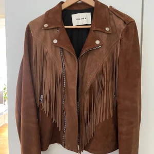 Brun mockajacka med fransar från BLK DNM - Säljer en brun mockajacka från BLK DNM med snygga fransar både fram och bak. Jackan har dragkedja, silvriga knappar och klassisk bikerstil. Perfekt för dig som vill sticka ut med en cool western-vibe. Passar till många olika outfits!