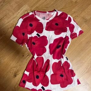 Vit t-shirt med röda blommor - Snygg vit t-shirt med stora röda blommor och svarta detaljer. T-shirten har rund hals och korta ärmar, perfekt för dig som gillar färgstarka prints. Materialet är mjuk bomull som känns skönt mot huden. Blommönstret ger en cool och lekfull vibe. Nytt skick!