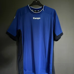 Blå fotbollströja från Kempa XL - Snygg blå tröja från Kempa i storlek XL. Tröjan har svarta detaljer på sidorna och ärmarna, rund halsringning med vit kant och tryckt Kempa-logga på bröstet. Tillverkad i lätt och funktionellt material som passar perfekt till träning eller match. Tröjan är oanvänd då jag beställde fel storlek