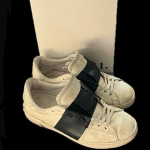 Valentino Garavani Open sneakers vit/svart - Snygga Valentino Garavani Open sneakers i vitt skinn med bred svart läderdetalj över vristen. Klassisk rund tå, vita snören och platt sula. Kommer med originalkartong och dustbag. Perfekta för dig som gillar lyxiga och stilrena sneakers.