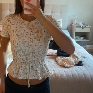 Zara Vit prickig blus med knyt i midjan - Supersöt vit blus med svarta prickar och korta ärmar. Toppen har en markerad midja med knytband och volang nertill. 