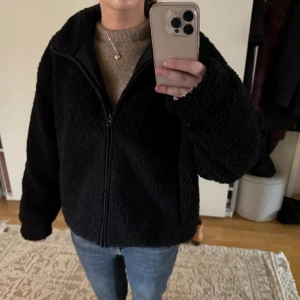 Svart teddyjacka med dragkedja - Mysig svart teddyjacka med dragkedja framtill och hög krage. Jackan har en loose passform och är tillverkad i fluffigt teddy-material som håller dig varm. Perfekt att slänga på sig över en hoodie eller stickad tröja. Enkel och stilren design som funkar till det mesta.