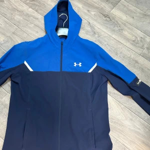Blå vindjacka från Under Armour - Snygg blå och marinblå vindjacka från Under Armour med huva och hel dragkedja. Jackan har vita detaljer, logga på bröstet. Sällsynt, finns inte i sverige. Köpt i Tjeckien. Priset kan diskuteras!!