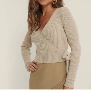 Beige ribbad omlottkofta Nakd - Superfin beige ribbad omlottkofta från NA-KD med knyt i sidan och v-ringad hals. Stickad i mjukt material och har långa ärmar. 