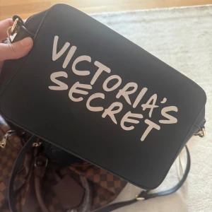 Svart axelväska - Cool svart axelväska med stort vitt tryck på framsidan. Väskan har en stilren, rektangulär form och en axelrem med gulddetaljer. Perfekt för att bära dina viktigaste grejer och ge din outfit en trendig touch.