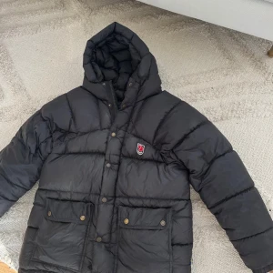 Svart dunjacka från Fjällräven XXXS - Svart dunjacka från Fjällräven med huva och stora fickor framtill. Jackan har Fjällrävens klassiska rävlogga på bröstet och stängs med både dragkedja och tryckknappar. Yttertyg och foder i polyamid, fyllning av 90% dun. Perfekt för kalla dagar. 