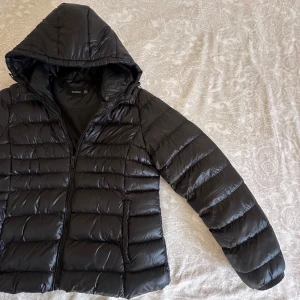 Svart pufferjacka med huva från Boohoo - Svart pufferjacka från Boohoo med glansig finish och huva. Jackan har horisontella quiltade partier, dragkedja framtill och långa ärmar. Perfekt för kalla dagar när du vill hålla stilen enkel och clean.