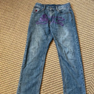 Ice cream byxor från billioner boys club  - Säljer ett par blåa jeans med coolt lila kattmotiv framtill och broderade bokstäver 'I' och 'C' i lila på bakfickorna. Jeansen har rak passform, klassisk femficksdesign och ett djurmönstrat patch vid linningen. Perfekta för dig som vill sticka ut.