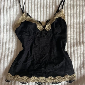 Svart spetslinnetopp med gulddetaljer - Super stylish black linen top with thin shoulder straps and decorative gold-colored lace edges at the top and bottom. The top has lace detail in the fabric and small buttons at the front. Perfect for those who like a chic and feminine style with extra details.
