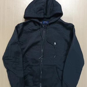 Svart hoodie från Polo Ralph Lauren - Svart hoodie från Polo Ralph Lauren med dragkedja framtill och klassisk huva. Tröjan har långa ärmar, två fickor och den ikoniska broderade loggan på bröstet. Tillverkad i mjuk bomull som är skön att bära. Perfekt för en chill och stilren look.