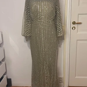 Festklänning i mintgrön glittrig långklänning - Säljer en elegant mintgrön långklänning med glittriga paljetter och ett rutigt mönster. Klänningen har långärmade, lätt puffiga ärmar och markerad midja. Materialet är skimrande och ger en lyxig känsla, perfekt för bal eller fest. Dragkedja bak för enkel påklädning.