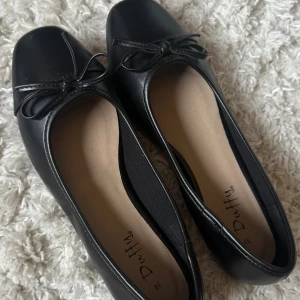 Svarta ballerinaskor från Duffy - Klassiska svarta ballerinaskor från Duffy med rund tå och söt rosettdetalj framtill. Skorna har en stilren design i syntetmaterial och passar perfekt till både jeans och klänning. Enkla att matcha och superbekväma för vardagen. Ej använda! 