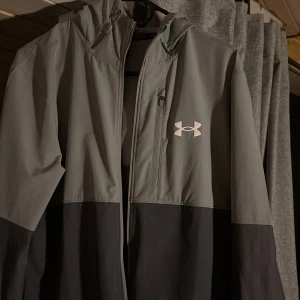 Under Armour vindjacka grå/mörkgrå - Snygg vindjacka från Under Armour i grått och svart med huva och dragkedja framtill. Jackan har en bröstficka med dragkedja och stor logga på bröstet. Perfekt för dig som gillar sportig stil och vill ha något lätt och funktionellt.