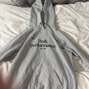 Grå hoodie från Peak Performance L - Snygg grå hoodie från Peak Performance med svart broderad logga på bröstet. Klassisk modell med huva, snörning och stor känguruficka framtill. Tillverkad i mjuk bomullsmix, perfekt för chill eller träning. Passar dig som gillar enkel och stilren streetwear. 
