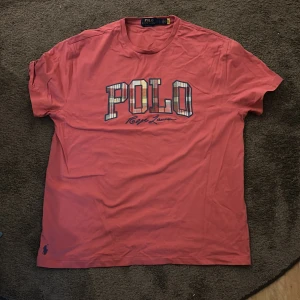 Rosa t-shirt från Polo Ralph Lauren - Snygg rosa t-shirt från Polo Ralph Lauren med stort tryck på bröstet där det står POLO i rutigt mönster och Ralph Lauren-signatur under. Classic fit och liten broderad logga nere till vänster. Priset kan diskuteras!!