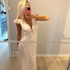 Vit långklänning från Nakd med blommigt mönster - Supersöt långklänning i vit med små rosa blommor, volangärmar och v-ringning med spetskant och knyt framtill. Klänningen har en romantisk vibe och är perfekt för varma dagar. Lätt och luftig modell som ger en avslappnad känsla.