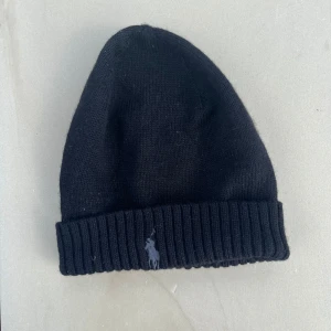 Svart stickad mössa från Polo Ralph Lauren - Svart mössa från Polo Ralph Lauren med klassisk ribbad kant och broderad logga framtill. Snygg och enkel design som passar till allt, stickad i mjukt material som håller dig varm. Perfekt accessoar för kyliga dagar.