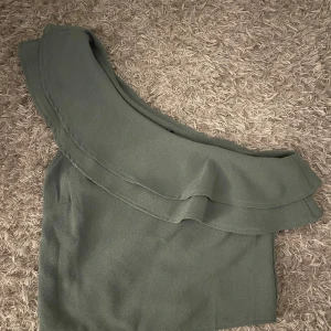 Grön oneshoulder topp Rut&Circle - Snygg grön topp från Rut&Circle i oneshoulder-modell med bred volang längs halsringningen. Toppen har en stilren och modern look, perfekt för dig som vill sticka ut lite extra. Materialet känns lätt och har en diskret struktur.