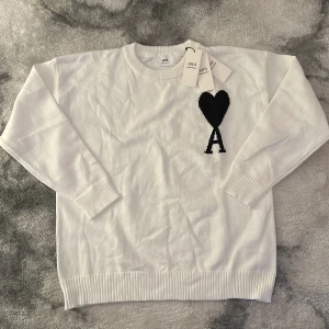 Vit stickad tröja från Ami Paris - Vit stickad tröja från Ami Paris med svart hjärta och bokstaven A broderat på bröstet. Tröjan har rund halsringning, ribbade muddar och lång ärm. Perfekt för dig som gillar stilrena och trendiga plagg med en twist.