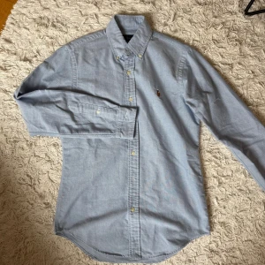 Ljusblå skjorta från Ralph Lauren XS - Klassisk ljusblå skjorta från Ralph Lauren i bomull. Skjortan har button-down krage, långa ärmar och knappar framtill. Broderad logga på bröstet ger en clean och tidlös look.