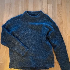 Mörkgrå stickad tröja från 157 - Mysig mörkgrå stickad tröja från 157 med rak passform och högre rund halsringning. Tröjan har långa ärmar och är perfekt för kyliga dagar. Materialet är fluffigt och ger en avslappnad vibe, lätt att styla med jeans eller kjol.