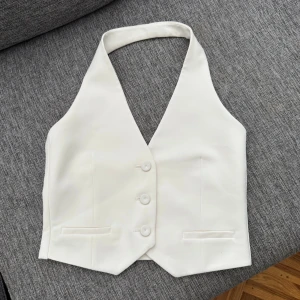 Vit omlottväst från Mango - Snygg vit väst från MNG med omlott-design och djup v-ringning. Västen har tre knappar framtill, två fejkade fickor och en stilren, figurnära passform. Den är aldrig använd! 