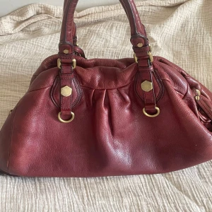 Vinröd väska, Marc by Marc Jacobs - Säljer en väldigt snygg vinröd väska i skinn från Marc by Marc Jacobs. Väskan har snygga guldfärgade detaljer, dubbla handtag och justerbar axelrem. På insidan finns ett fiskbensmönstrat foder och ett dragkedjefack samt två fickor. Perfekt för dig som vill ha en rymlig och stilig väska! Bredd: 42 cm, Djup: 21 cm, Höjd: 18 cm