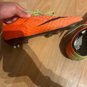 Nike orange fotbollsskor med snörning, hypervenom - Säljer ett par Nike fotbollsskor i orange med svarta detaljer och gröna snören. Skorna har en texturerad ovandel i syntetmaterial och är designade för bra grepp på planen. Perfekta för dig som vill sticka ut på matchen!