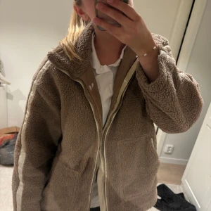 Beige teddyjacka med dragkedja - Mysig beige teddyjacka med vit dragkedja och vita detaljer längs ärmarna. Jackan har hög krage, två stora fickor framtill och en loose passform som ger en chill vibe. Perfekt för kalla dagar när du vill vara både varm och snygg. Säljer då den inte satt som jag ville. Köpt här från Plick så vet tyvärr inte märke. Öppen för att diskutera pris 