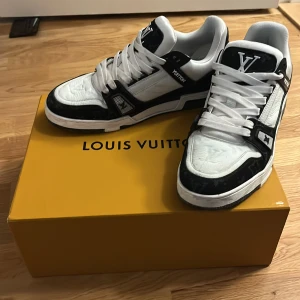 Louis Vuitton svartvita sneakers - Säljer ett par Louis Vuitton sneakers i svart och vitt med tydliga LV-loggor på både plös och häl. Skorna har snörning, platt sula och är tillverkade i skinn med detaljer i syntet. Klassisk och lyxig design med kontrasterande färger och diskreta monogramdetaljer.
