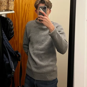 Grå stickad tröja från Sandö - Mysig grå stickad tröja från Sandö i storlek M. Tröjan har en klassisk rund halsringning, ribbade muddar och nederkant. Perfekt för lager-på-lager och chill dagar. Enkel och stilren design som funkar till jeans eller chinos.