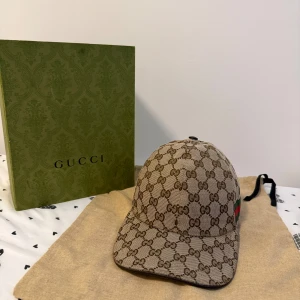 Gucci beige keps med GG-mönster - Snygg beige keps från Gucci med det klassiska GG-monogrammet över hela kepsen. Kepsen har en böjd skärm och justerbar rem baktill. Materialet är bomull och polyester, och på sidan finns Guccis ikoniska gröna och röda ränder. Perfekt för dig som vill ha en lyxig streetstyle. Box och dustbag följer med! Vid frågor eller funderingar så är det bara att höra av sig!!
