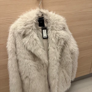 Pälsjacka (fuskpäls)  - Säljer en pälsjacka (fuskpäls) från lager 157. Vit/grå/beige färg med krage, fickor och foder inuti. Jackan knäpps med två ”osynliga” knappar. Varm och skön jacka, perfekt att ha just nu🤩. Helt ny med lappar kvar, strl XS/S (normal/lite oversize passform). Nypris 600, säljer för 250🩶