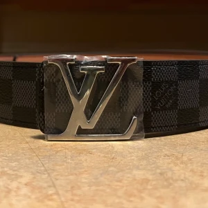 Louis Vuitton bälte - Snyggt svart bälte från Louis Vuitton med klassiskt rutigt mönster och en stor silverfärgad LV-logga som spänne. Bältet är gjort i skinn och har en stilren, modern look som verkligen sticker ut. Perfekt för dig som vill ha något exklusivt och trendigt.