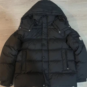 Svart Moncler pufferjacka ( MED KVITTO ) - Säljer en svart pufferjacka från Moncler med stor huva och feta, quiltade sektioner. Jackan har Moncler-logga på ärmen, praktiska fickor och både knappar och dragkedja framtill. Perfekt för dig som vill ha en stilren och varm jacka. Jackan är perfekt till vintern. 10 utav 10 en super fet och unik jacka. Kvitto finns, bara att fråga om ni vill se.