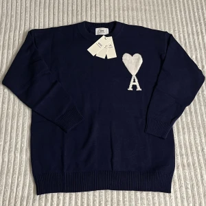 Ami Paris Tröja - Snygg mörkblå stickad tröja från Ami Alexandre Mattiussi med vit hjärtlogga och bokstaven A på bröstet. Tröjan har rund hals, ribbade muddar och är långärmad. Perfekt för dig som gillar stilrena och trendiga plagg. Tröjan är mjuk och kön och passar perfekt inför nuvarande vinter fast även kommande vår och sommar. Ett till ett. Tröja säljs på grund av fel storlek. Vid några funderingar är det bara att skriva. 