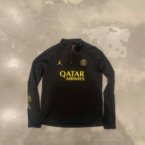 PSG x Jordan svart half zip tröja - Storlek 158-170cm. Svart PSG x Jordan långärmad tröja med gul logga och detaljer, half zip framtill och Paris-tryck på ärmen. Slim fit-modell i mjukt syntetmaterial, perfekt för fotbollsträning eller chill. Snyggt mönster på ärmarna och klassisk Jordan-logga på bröstet.