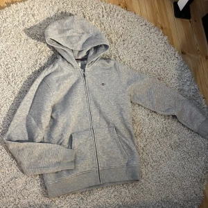 Grå hoodie från Gant med dragkedja - Snygg ljusgrå hoodie från Gant med broderad logga på bröstet. Tröjan har huva, dragkedja framtill och två fickor. Tillverkad i mjuk bomullsmix och har ribbade muddar vid ärmslut och nederkant. Perfekt för chill dagar eller när du vill vara casual.