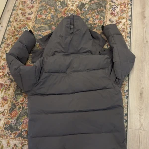 Mörkgrå dunjacka från Canada Goose - Säljer en mörkgrå dunjacka från Canada Goose med huva och klassisk logga på ärmen. Jackan har långa ärmar, dragkedja och knappar framtill, samt stora fickor. Perfekt för kalla dagar och har en schysst oversized look.