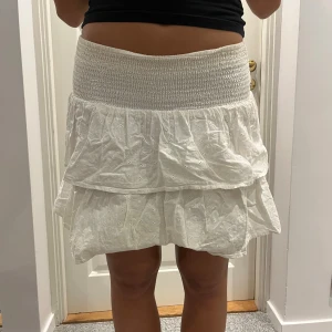 Vit volangkjol med broderade detaljer - Supersöt vit kortkjol med bred smock i midjan och dubbla volanger. Kjolen har broderade små hålmönster som ger en romantisk känsla. Perfekt för varma dagar och lätt att matcha med en enkel topp.