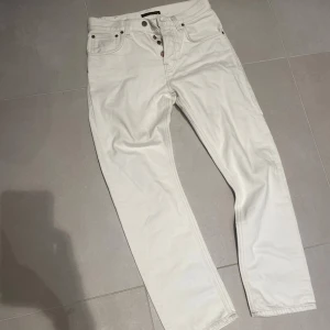 Vita jeans från Nudie Jeans - Snygga vita jeans från Nudie Jeans med klassisk femficksdesign och raka ben. Byxorna har knäppning med flera knappar framtill och subtila detaljer på bakfickorna. Perfekt för dig som gillar stilrena och fräscha jeans.