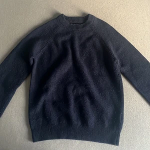 Mörkblå stickad tröja i lammull - Mysig mörkblå stickad tröja från C i lammullsmix. Tröjan har rund hals och långa ärmar med ribbade muddar vid hals, ärmslut och nederkant. Perfekt för dig som gillar klassisk och enkel stil med en soft vibe.